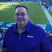 A man in a purple shirt standing in front of a stadium.